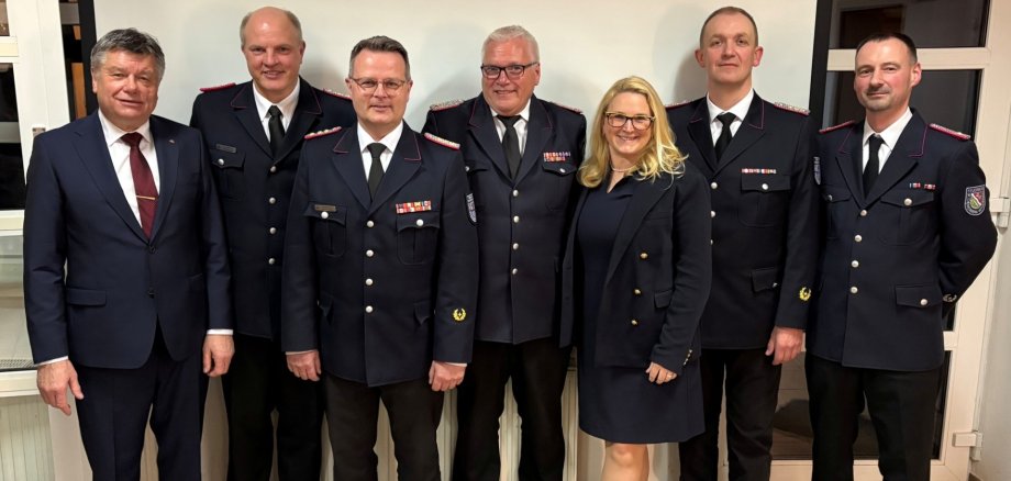 Sieben Personen stehen nebeneinander vor einer Leinwand in einem Innenraum. Sechs Männer tragen Feuerwehruniformen mit Abzeichen, eine Frau trägt ein dunkles Kostüm. Die Gruppe zeigt die aktuelle und ehemalige Amtswehrführung des Amtes Sandesneben-Nusse.