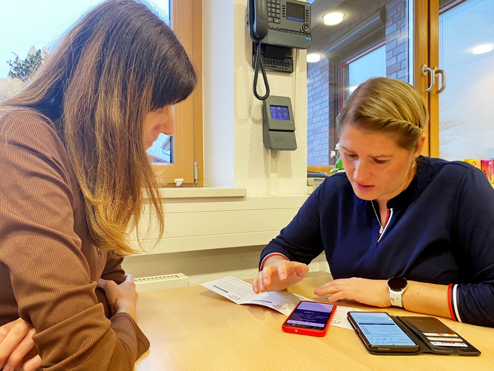 Zwei Frauen sitzen an einem Tisch und betrachten gemeinsam ein Dokument. Eine Person zeigt auf den Inhalt, während auf dem Tisch zwei Smartphones und weitere Unterlagen liegen. Im Hintergrund sind Fenster und technische Geräte an der Wand zu sehen.