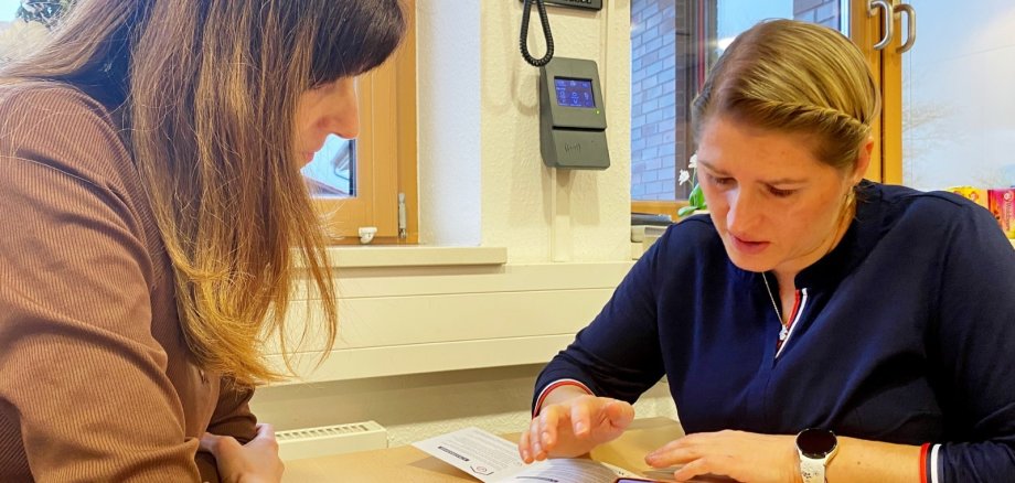 Zwei Frauen sitzen an einem Tisch und betrachten gemeinsam ein Dokument. Eine Person zeigt auf den Inhalt, während auf dem Tisch zwei Smartphones und weitere Unterlagen liegen. Im Hintergrund sind Fenster und technische Geräte an der Wand zu sehen.