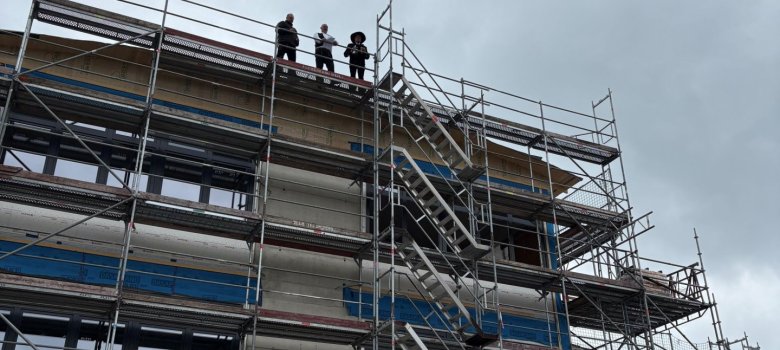 Mehrgeschossiger Rohbau mit Baugerüst während eines Richtfestes. Über dem Gebäude hängt eine Richtkrone an einem Baukran. Auf dem Gerüst stehen mehrere Personen unter grauem Himmel.