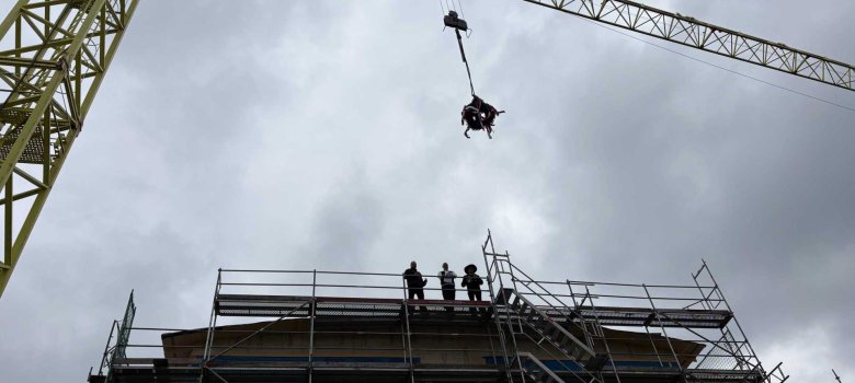 Auf einer Baustelle hängt eine Richtkrone an einem Baukran über einem mehrgeschossigen Gebäude im Rohbau. Mehrere Personen stehen auf dem Baugerüst und beobachten das Richtfest bei bewölktem Himmel.