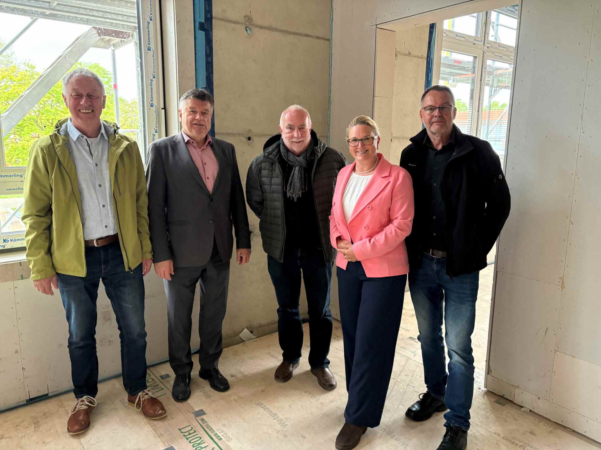 Fünf Personen stehen für ein Gruppenfoto in den Innenräumen eines im Bau befindlichen Gebäudes. Im Hintergrund sind unverkleidete Wände, Fenster und Baustellenmaterialien zu sehen.