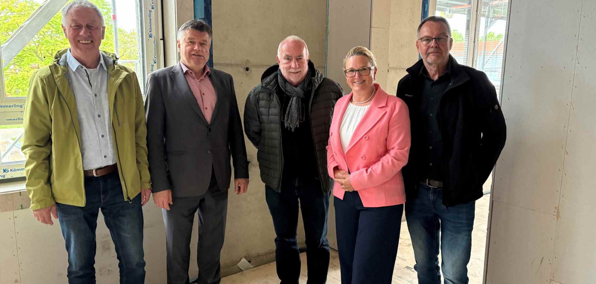Fünf Personen stehen für ein Gruppenfoto in den Innenräumen eines im Bau befindlichen Gebäudes. Im Hintergrund sind unverkleidete Wände, Fenster und Baustellenmaterialien zu sehen.