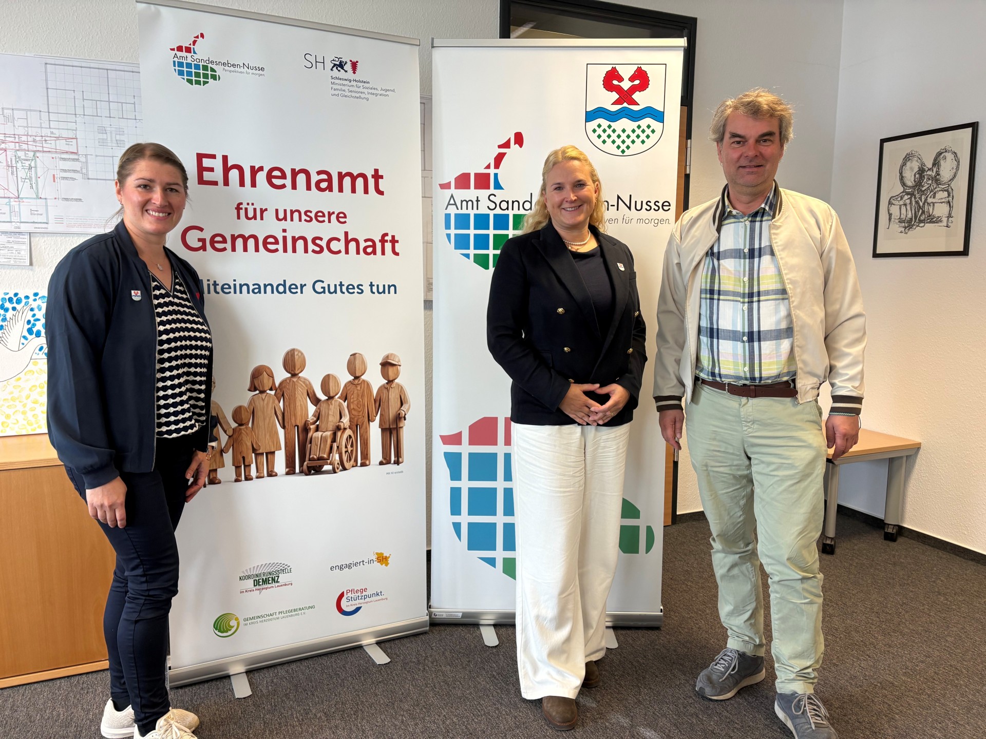 Tatjana Skowronski, Tina Knuth und Oliver Brandt stehen vor zwei Bannern zum Ehrenamt und des Amtes Sandesneben-Nusse.