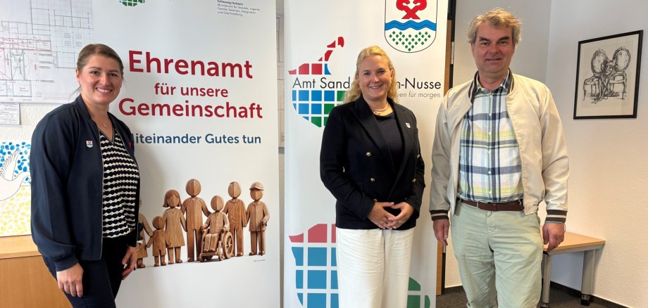 Tatjana Skowronski, Tina Knuth und Oliver Brandt stehen vor zwei Bannern zum Ehrenamt und des Amtes Sandesneben-Nusse.