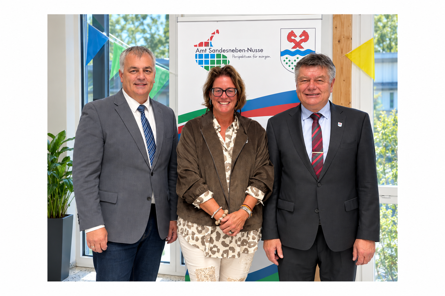 ars Wunsch, 1. stellvertretender Amtsvorsteher, Nicole Demir, 2. stellvertretende Amtsvorsteherin, und Amtsvorsteher Ulrich Hardtke stehen gemeinsam im Amtsgebäude vor dem Logo des Amtes Sandesneben-Nusse.