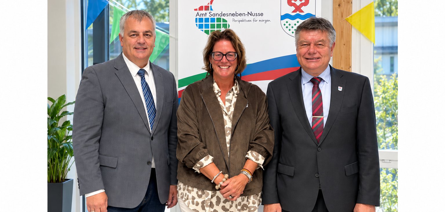 ars Wunsch, 1. stellvertretender Amtsvorsteher, Nicole Demir, 2. stellvertretende Amtsvorsteherin, und Amtsvorsteher Ulrich Hardtke stehen gemeinsam im Amtsgebäude vor dem Logo des Amtes Sandesneben-Nusse.