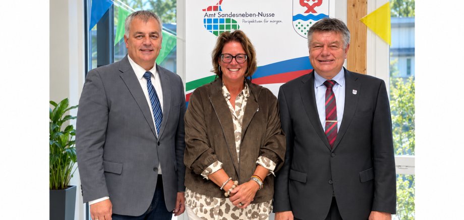 ars Wunsch, 1. stellvertretender Amtsvorsteher, Nicole Demir, 2. stellvertretende Amtsvorsteherin, und Amtsvorsteher Ulrich Hardtke stehen gemeinsam im Amtsgebäude vor dem Logo des Amtes Sandesneben-Nusse.