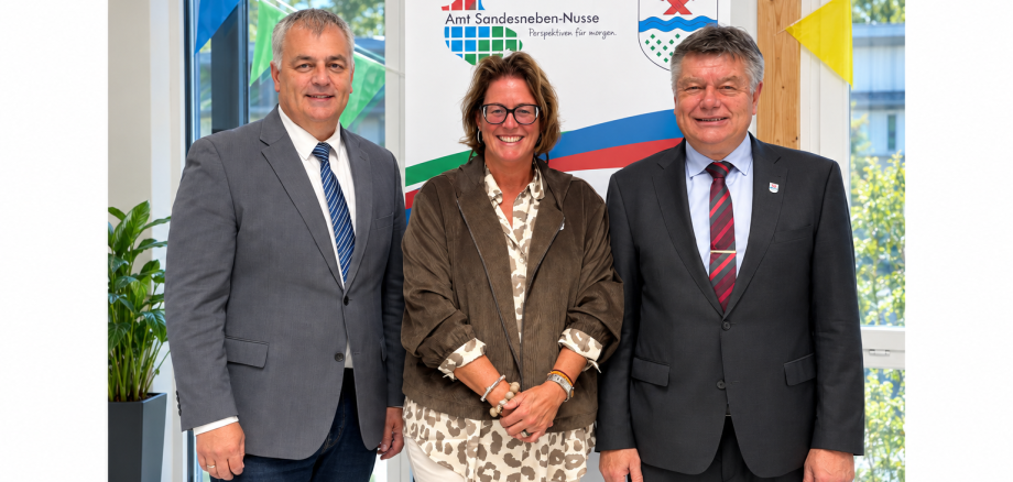 ars Wunsch, 1. stellvertretender Amtsvorsteher, Nicole Demir, 2. stellvertretende Amtsvorsteherin, und Amtsvorsteher Ulrich Hardtke stehen gemeinsam im Amtsgebäude vor dem Logo des Amtes Sandesneben-Nusse.