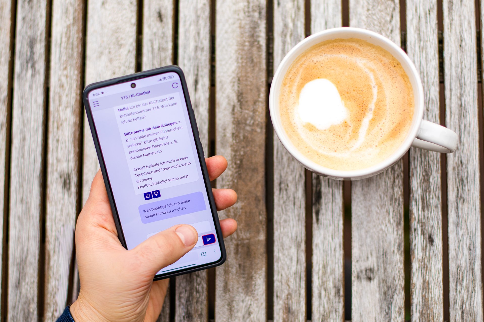 Eine Person hält ein Smartphone, auf dem der 115-KI-Chatbot geöffnet ist. Daneben steht eine Tasse Kaffee auf einem Holztisch. Das Bild symbolisiert den einfachen mobilen Zugang zu Verwaltungsinformationen über den Chatbot.
