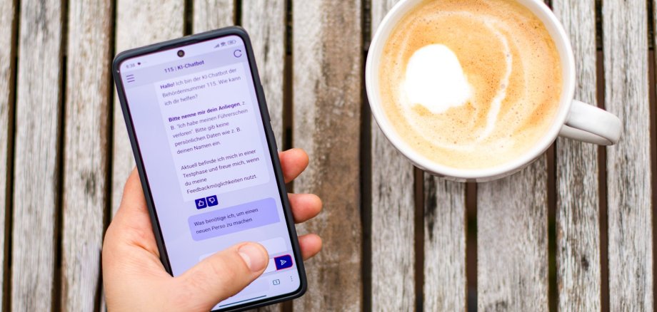 KI-generiertes Bild Eine Person hält ein Smartphone, auf dem der 115-KI-Chatbot geöffnet ist. Daneben steht eine Tasse Kaffee auf einem Holztisch. Das Bild symbolisiert den einfachen mobilen Zugang zu Verwaltungsinformationen über den Chatbot.