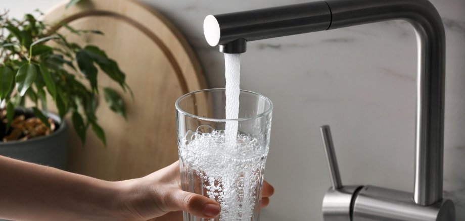Hand hält ein Glas unter einen Wasserhahn, aus dem frisches Leitungswasser läuft.
