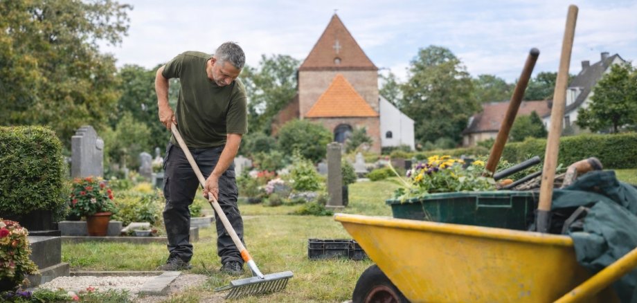 Ein Friedhofsgärtner arbeitet auf einer gepflegten Friedhofsanlage und harkt Kies auf einem Grab, während im Hintergrund eine Kirche und weitere Grabstätten zu sehen sind. Im Vordergrund steht eine Schubkarre mit Gartenwerkzeug und Blumen.