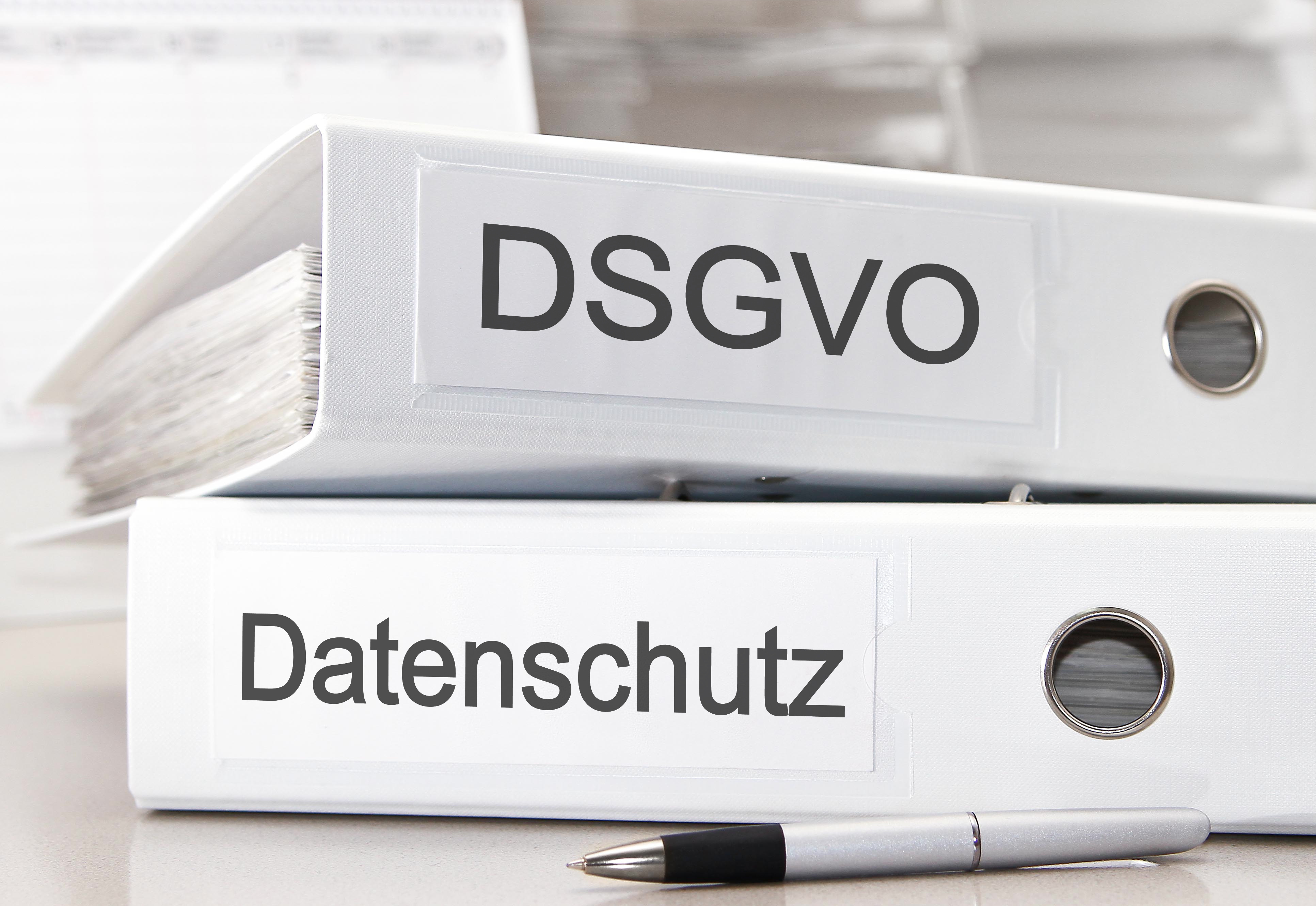 Zwei weiße liegende Aktenordner mit den Beschriftungen DSGVO und Datenschutz. Davor liegt ein silberner Kugelschreiber.