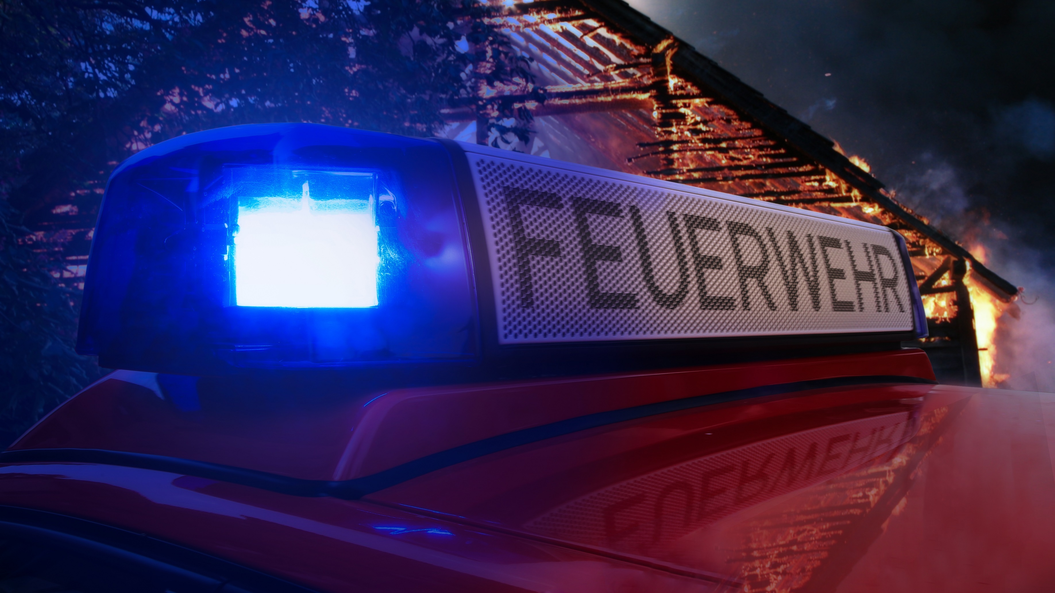 Feuerwehrfahrzeug mit eingeschaltetem Blaulicht vor einem brennenden Gebäude.