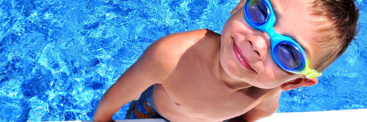 Ein lachender Junge mit Schwimmbrille hält sich am Rand eines klaren, blauen Pools fest. Er trägt eine Badehose und genießt sichtlich das Schwimmen an einem sonnigen Tag.