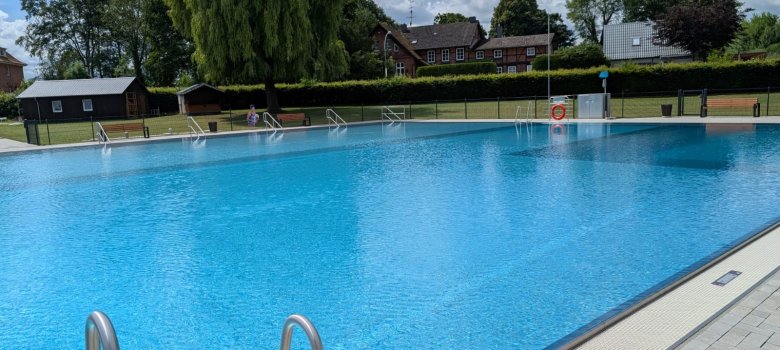 Großes Schwimmbecken im Freibad Steinhorst mit klarem, blauem Wasser, Edelstahlleitern und umliegenden Liegeflächen, im Hintergrund Bäume und Wohnhäuser.