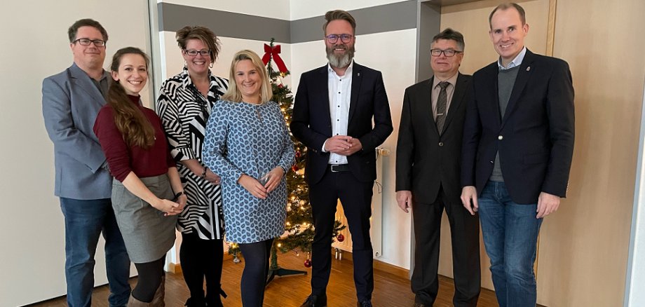 In dem Bild sind sieben Personen zu sehen, von links nach rechts: Herr Völkering, Frau Sülflohn, Frau Demir, Frau Knuth, Herr Madsen, Herr Hardtke, Herr Doktor Mager.