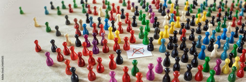 Viele bunte Spielfiguren sind in großen Gruppen auf einem Tisch angeordnet, die Farben reichen von Rot, Schwarz, Gelb, Blau bis Grün. In der Mitte befindet sich ein Stück Papier mit einem roten Kreuz, das an eine Wahlurne oder eine Abstimmung erinnert.