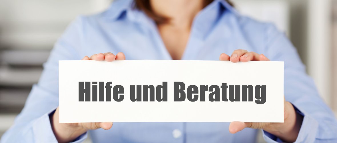 Person in blauem Hemd hält ein Schild mit der Aufschrift „Hilfe und Beratung“ in die Kamera – Symbolbild für Unterstützungs- und Beratungsangebote.