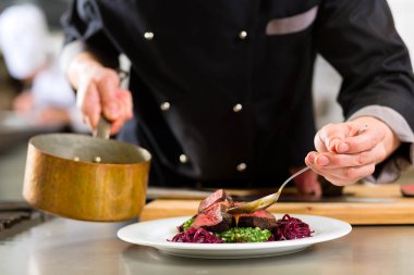 Koch in schwarzer Uniform richtet sorgfältig ein Gericht mit Fleisch, Rotkohl und Soße auf einem Teller an – Symbol für kulinarische Qualität und Handwerkskunst in der Gastronomie.