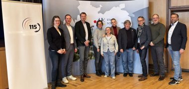 Gruppenfoto von zehn Personen, die in einem Veranstaltungsraum vor einem Roll-Up mit dem 115-Logo und einer Wandgrafik stehen.