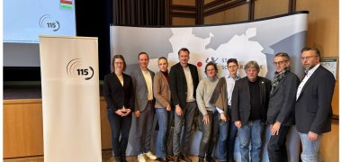 115.de Gruppenfoto von neun Personen, die in einem Veranstaltungsraum vor einem Roll-Up mit dem 115-Logo und einer Wandgrafik stehen.