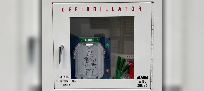 Defibrillator (AED) Wandkasten mit einem Defibrillator (AED) hinter einer Glasscheibe. Auf dem Gerät ist eine Zeichnung zu sehen, die die richtige Platzierung der Elektroden auf dem Oberkörper zeigt.