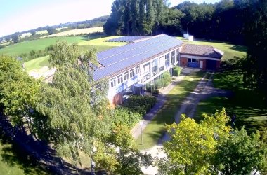 Luftaufnahme eines langgestreckten Gebäudes mit großer Solaranlage auf dem Dach, umgeben von Bäumen und Wiesen.