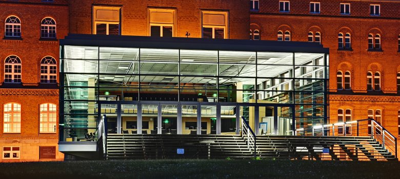 Das Foto zeigt das Landtagsgebäude des Landes Schleswig-Holstein in Kiel. Der klassizistische Backsteinbau wurde durch einen modernen Glasvorbau ergänzt, der abends hell erleuchtet ist und einen architektonischen Kontrast zwischen Tradition und Moderne bildet.